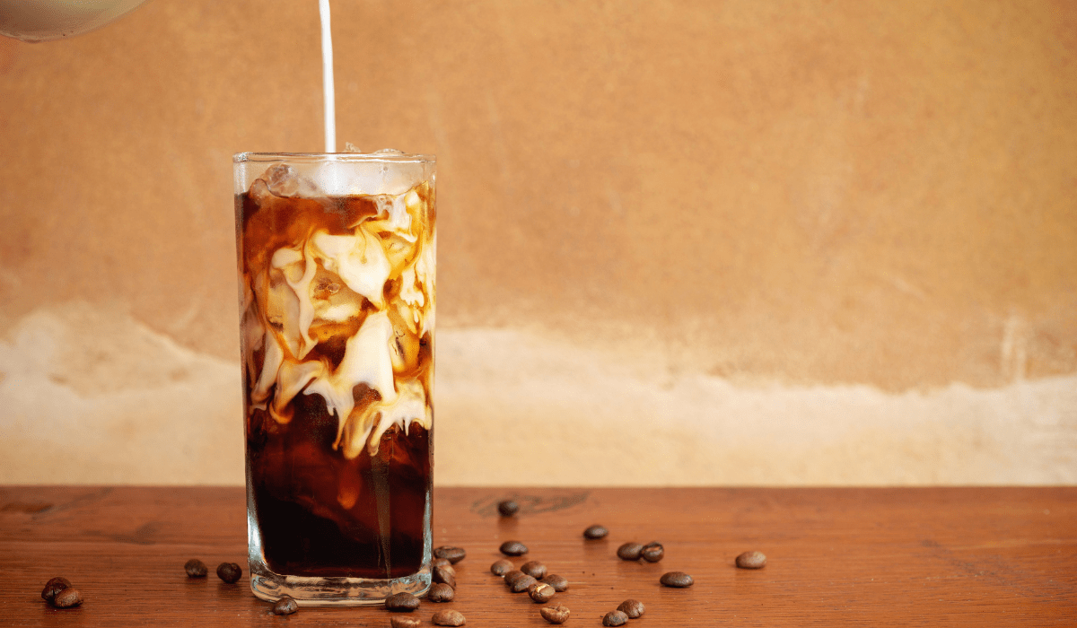 Cold brew coffee with ice and a splash of cream, served in a tall glass with coffee beans scattered around.