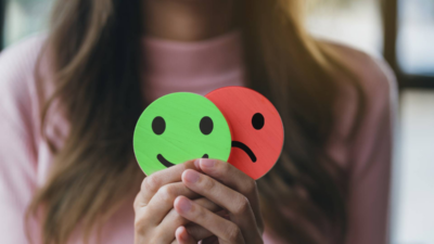 A woman holding two wooden emoji faces—one green and smiling, the other red and sad—symbolizing mood swings and mental health.