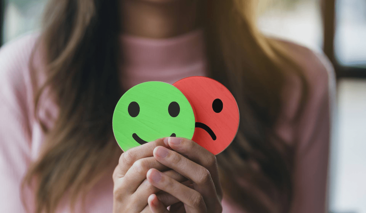 A woman holding two wooden emoji faces—one green and smiling, the other red and sad—symbolizing mood swings and mental health.