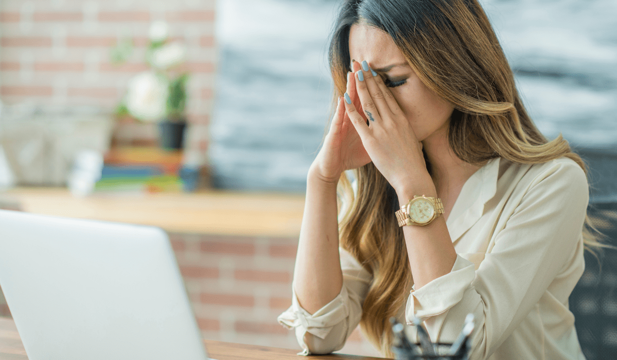 A stressed woman sitting at her desk with her hands on her face, experiencing workplace burnout and exhaustion.