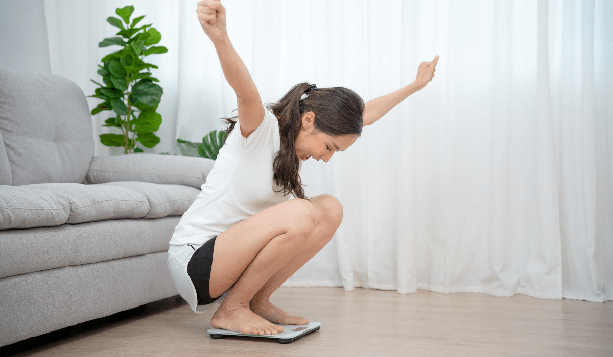 A happy woman celebrating her weight loss success after achieving her fitness goals with Beneve Xcelerate Coffee, standing on a scale at home.